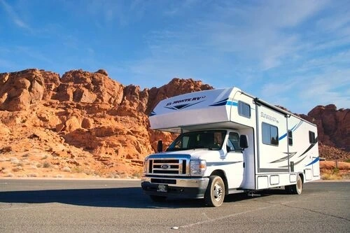 A white El Monte RV motorhome parked on asphalt in front of vibrant red rock formations under a bright blue sky.