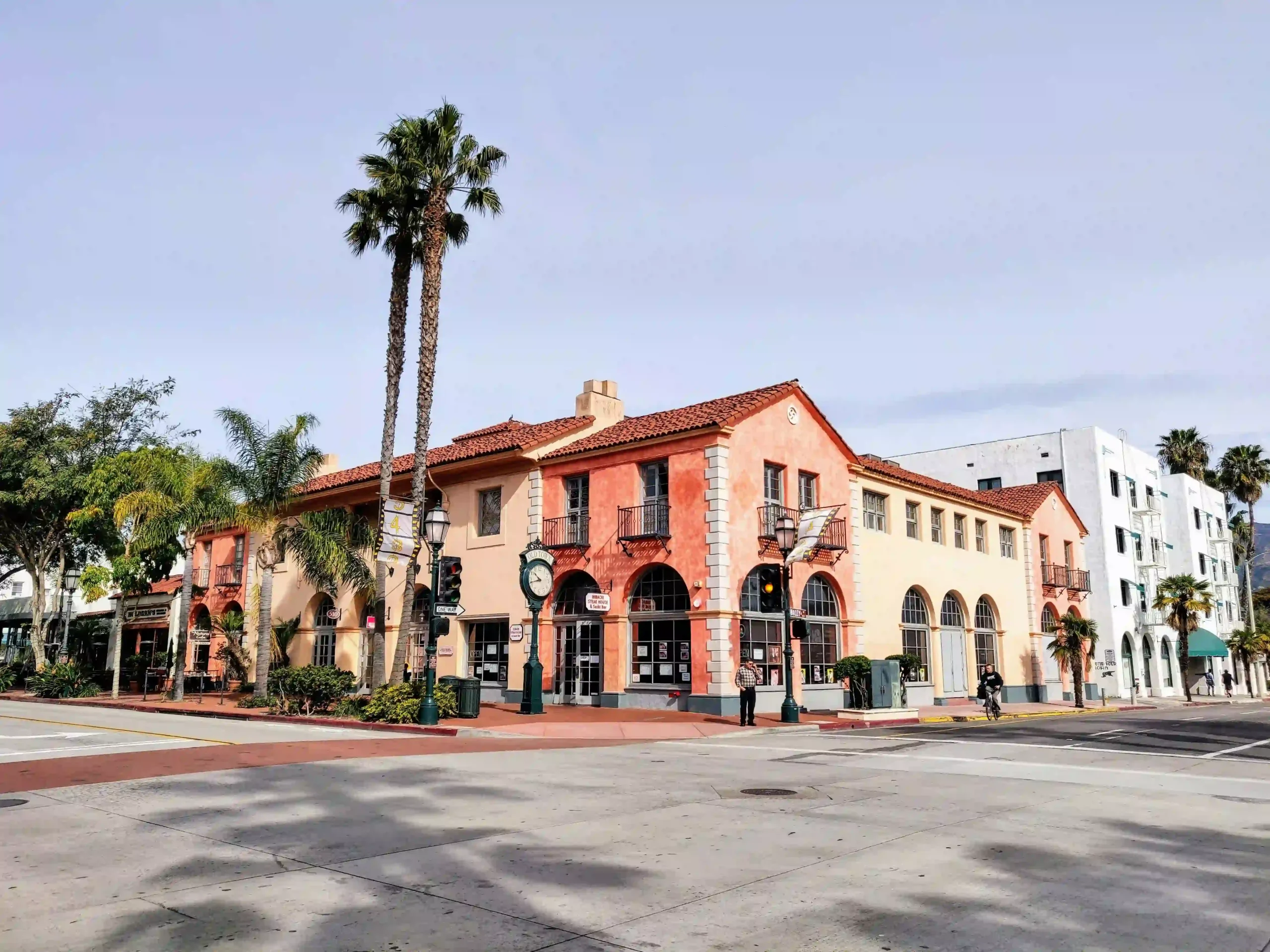 Mediterranean-style salmon and cream-colored buildings with red tile roofs and arched windows on a street corner in Santa Barbara, featuring tall palm trees.