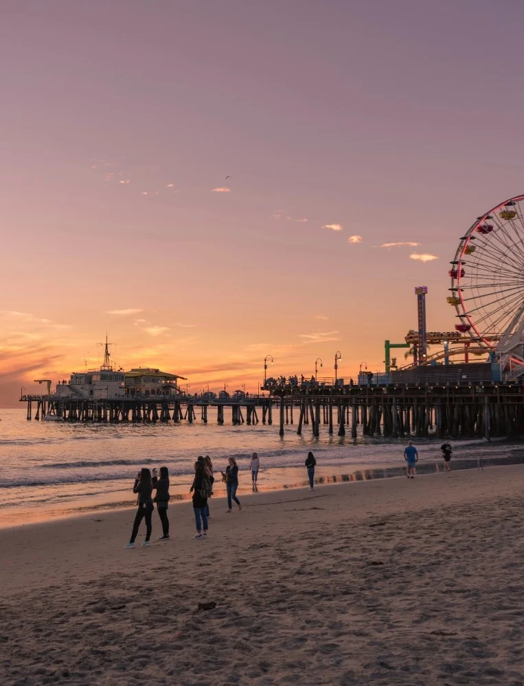 A wide sunset view of Santa Monica Pier and the Pacific Park Ferris wheel from the sandy beach, with silhouettes of people walking along the shoreline under a vibrant purple and orange sky.