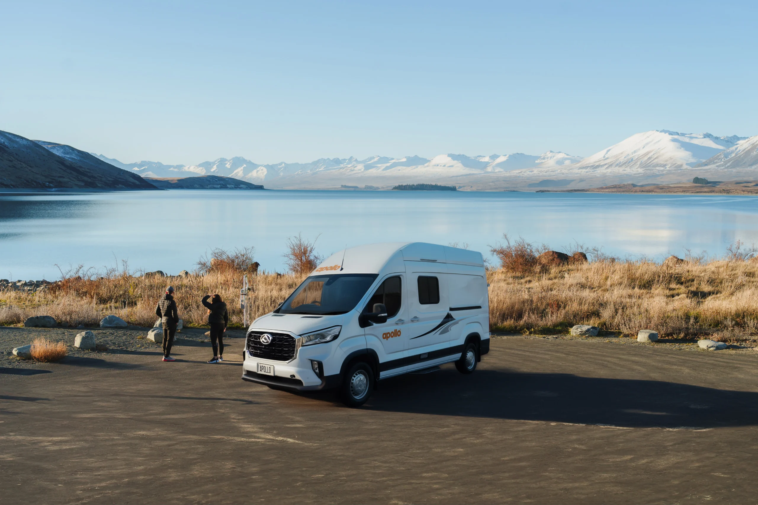 White Apollo camper parked overlooking a blue lake with snow-capped mountains and two travelers enjoying the view.