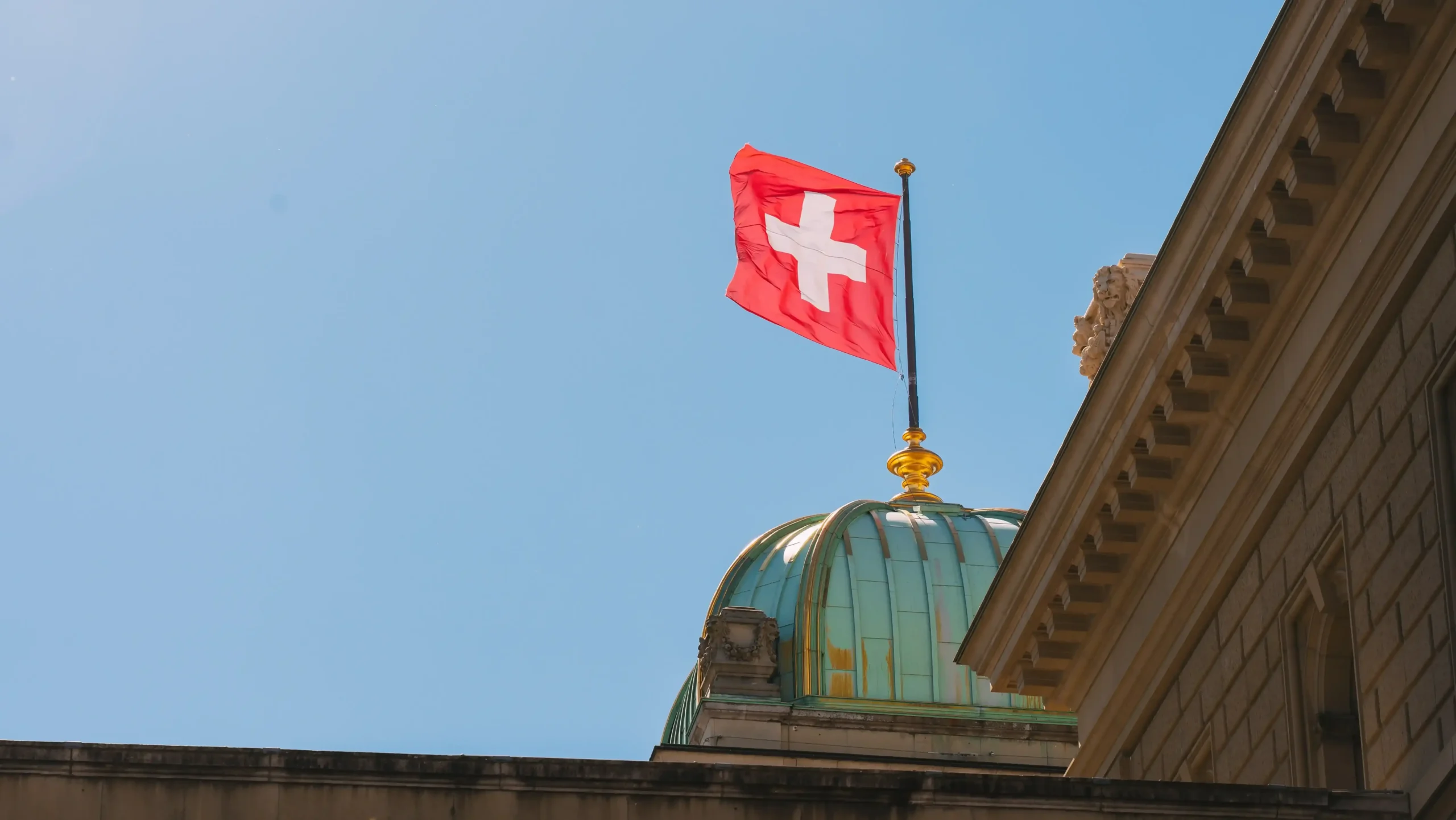 The Swiss national flag waving from the top of the green copper dome of the Federal Palace (Bundeshaus) in Bern under a bright blue sky.