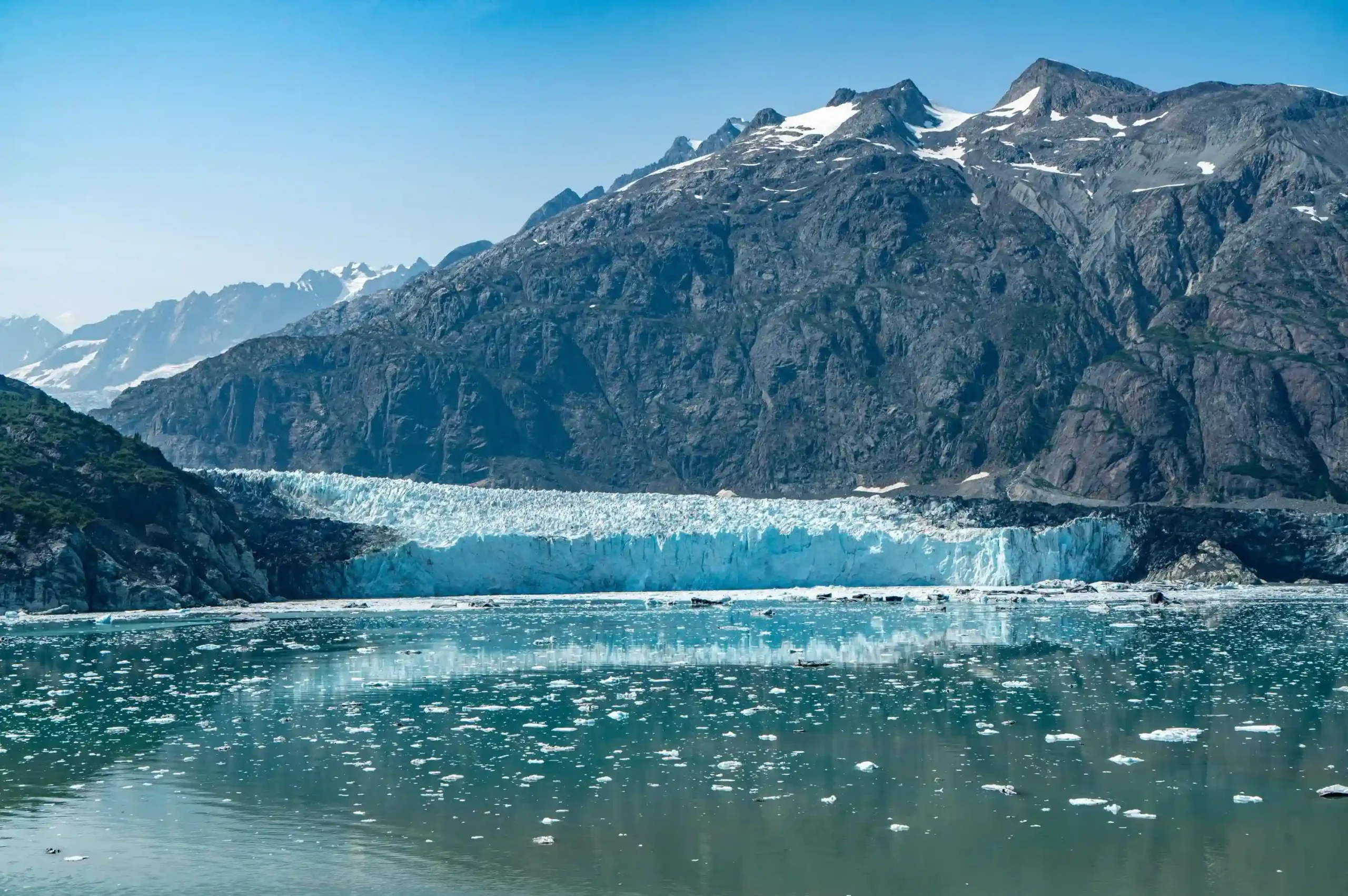 A massive blue-white glacier meeting the sea. Small icebergs and brash ice float in the calm, turquoise water in the foreground.