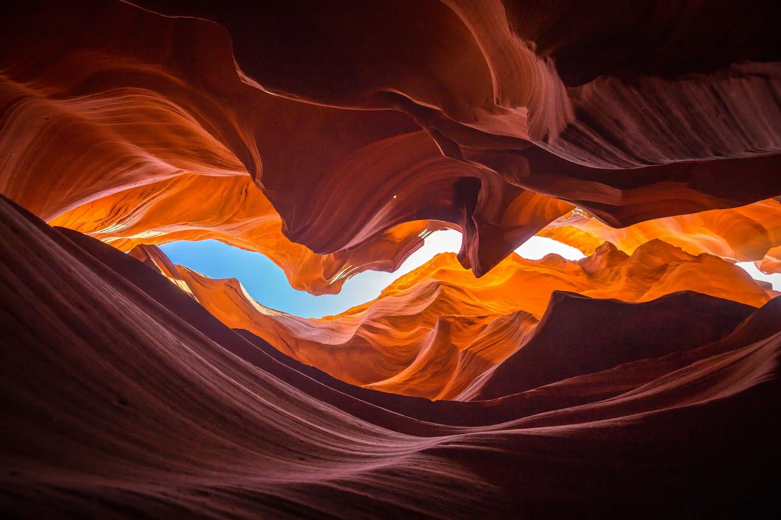 The flowing, sculpted sandstone walls in warm shades of orange and brown rise steeply, framing a narrow sliver of bright blue sky in the centre. The texture of the rock resembles frozen waves.
