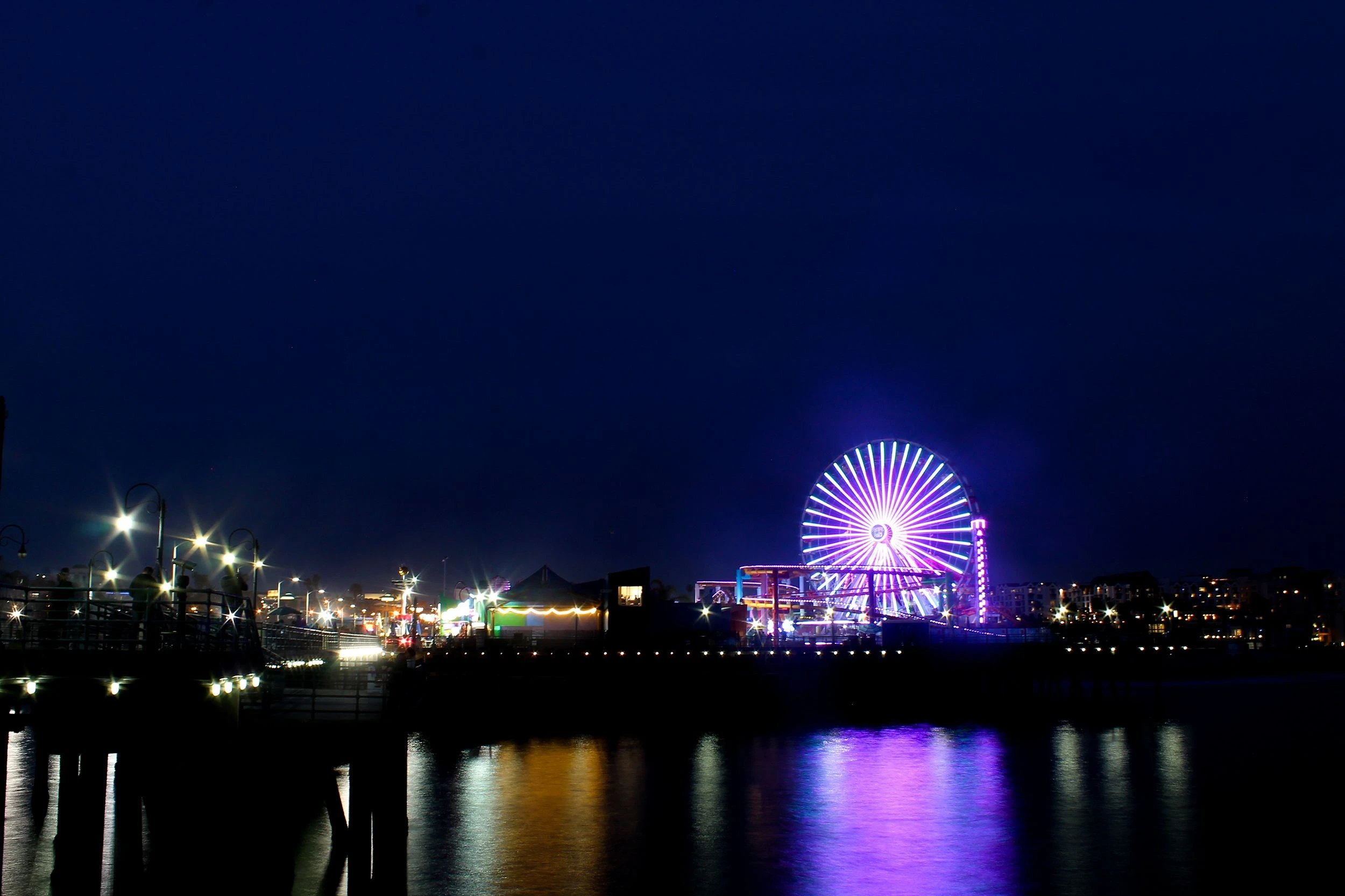 A night view of the Santa Monica Pier featuring a large, brightly lit purple and white Ferris wheel reflected in the dark water below.