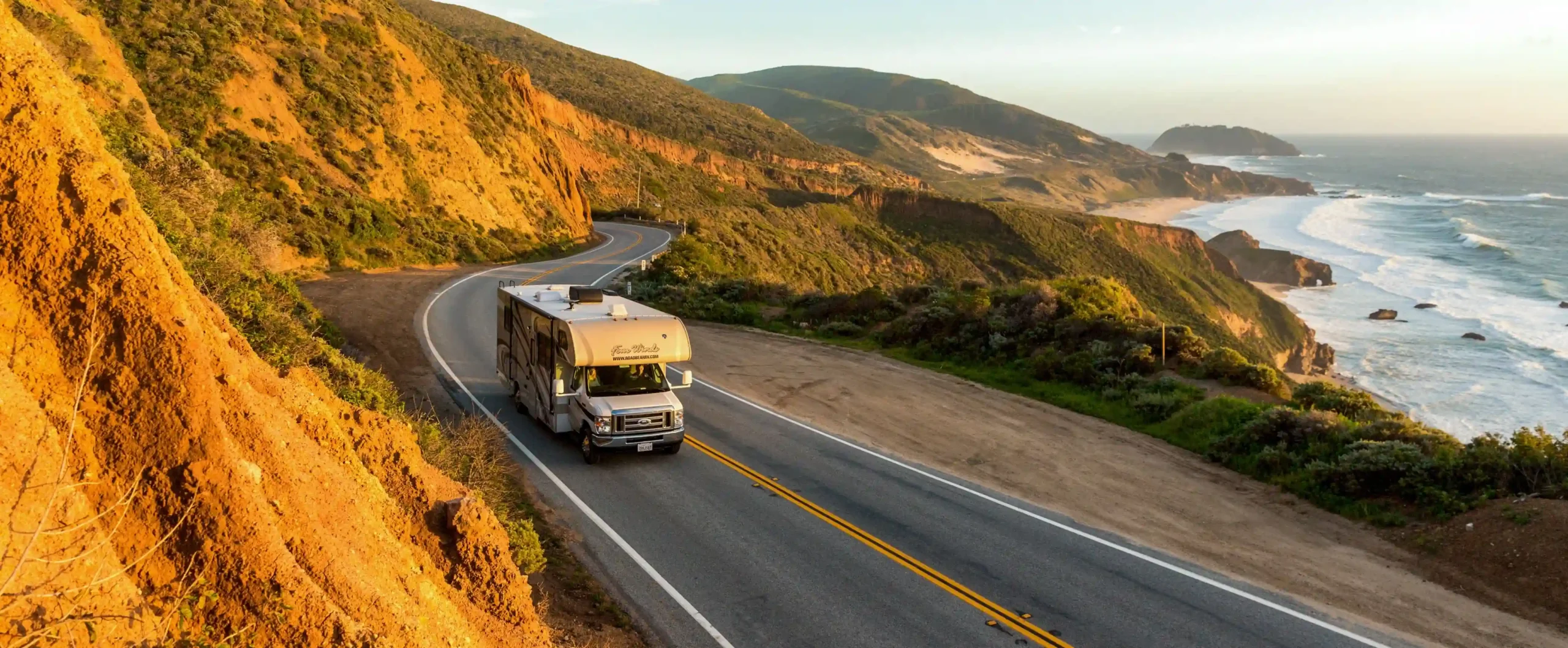 High-angle view of an RV driving on Highway 1 along steep coastal bluffs overlooking the Pacific Ocean at sunset.