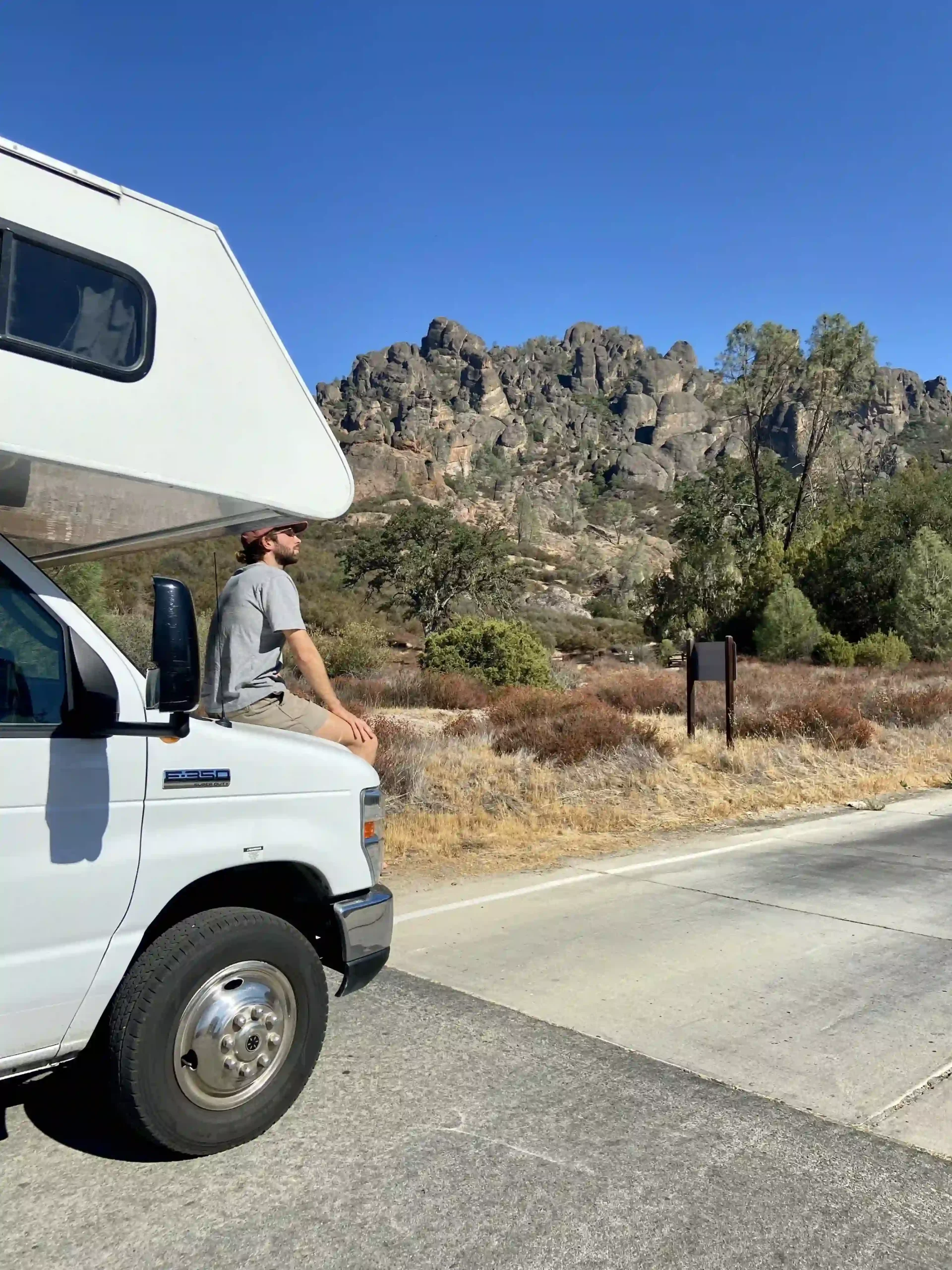 A man in a grey t-shirt sits on the hood of a white El Monte RV, looking toward jagged, rocky mountains under a clear blue sky.