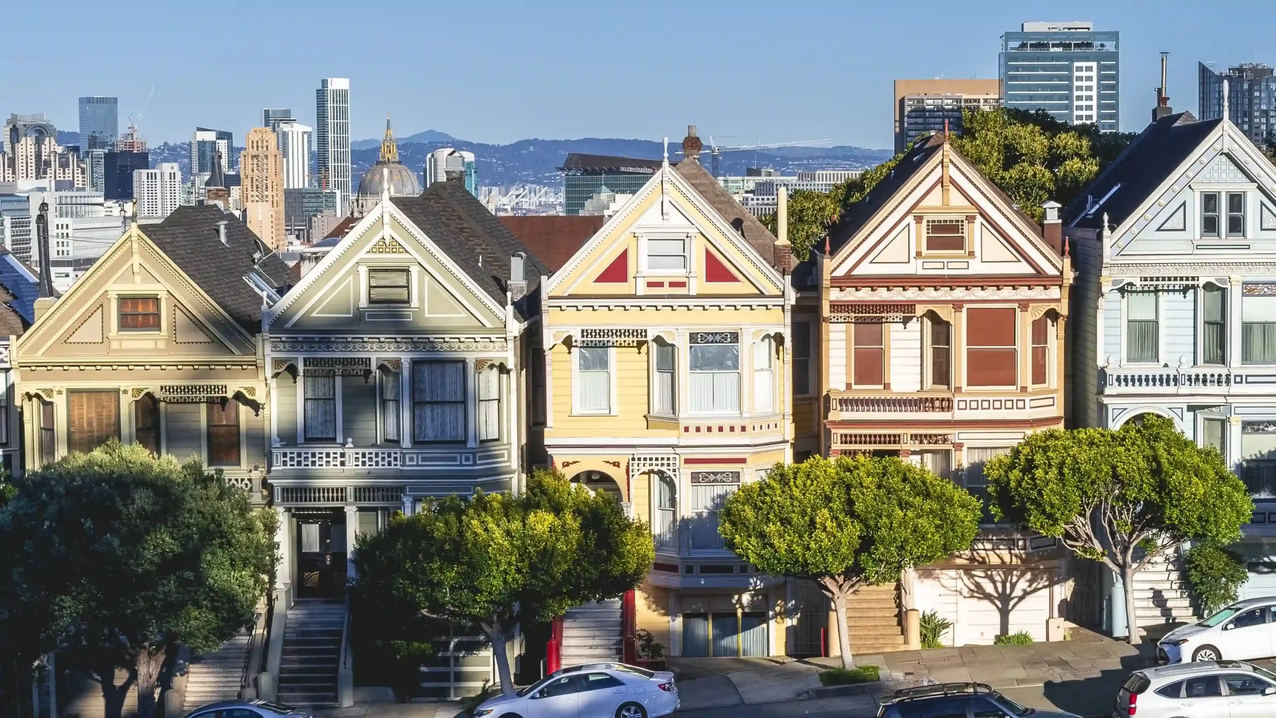 A row of Victorian timber houses stand on a sloping city street, with green leafy trees in front and the high-rise San Francisco downtown skyline rising in the background.