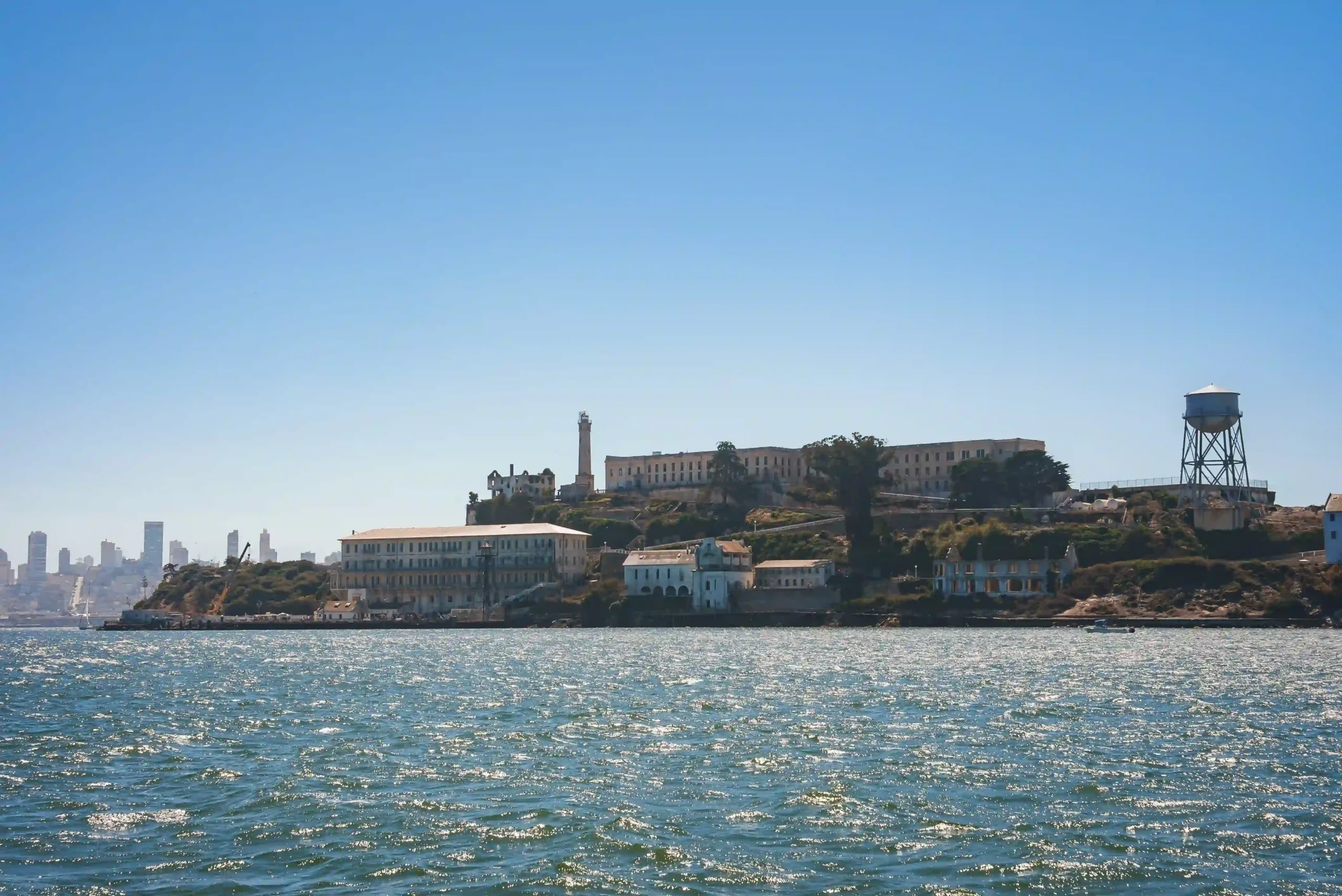 A wide shot across sparkling blue bay water toward Alcatraz Island, showing its historic cellhouse, lighthouse, and water tower on the rocky hilltop with the faint San Francisco skyline in the background.