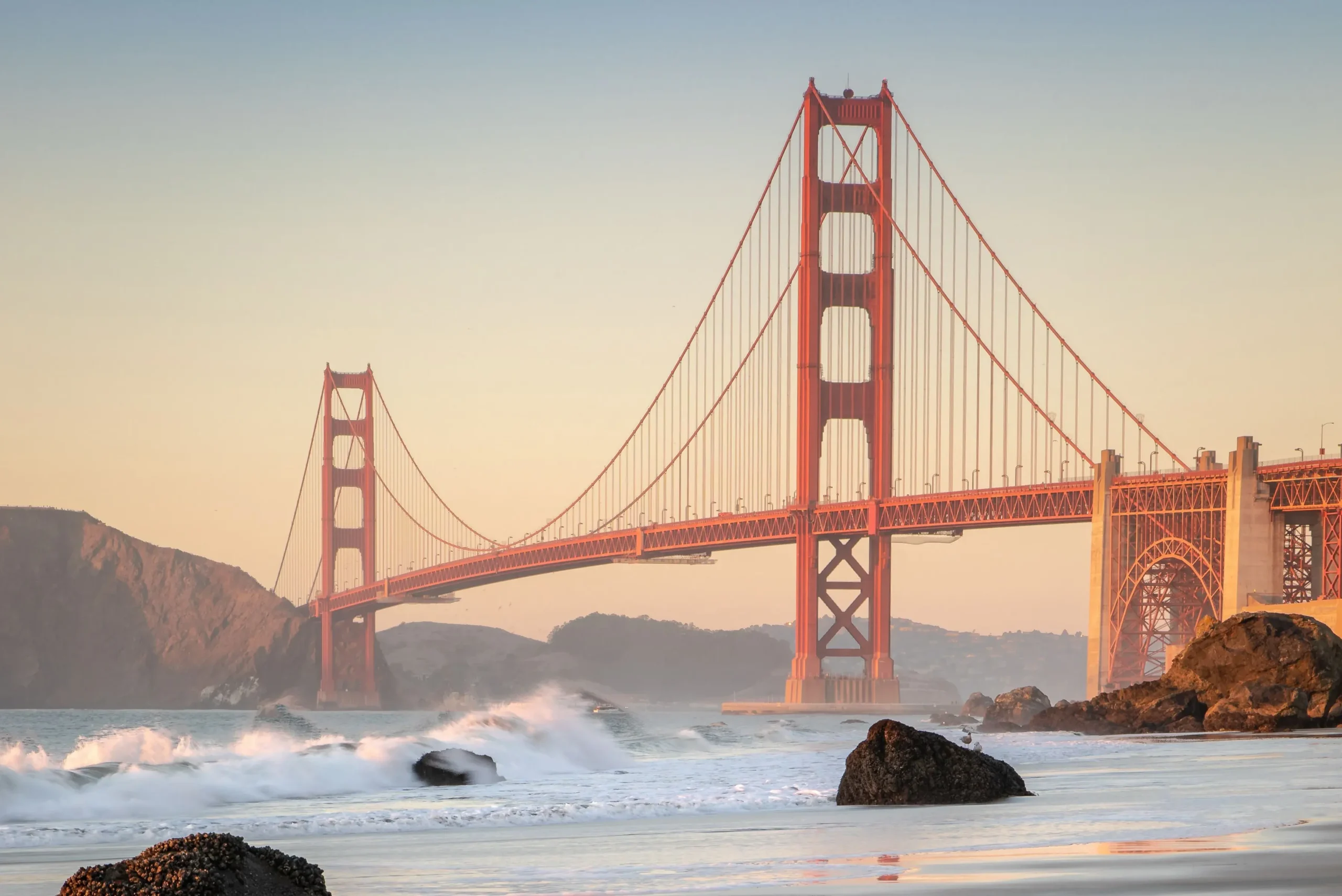 A wide view of the Golden Gate Bridge stretching across the frame, seen from the shoreline of Baker Beach as white ocean waves crash over dark rocks on the wet sand under soft golden light.
