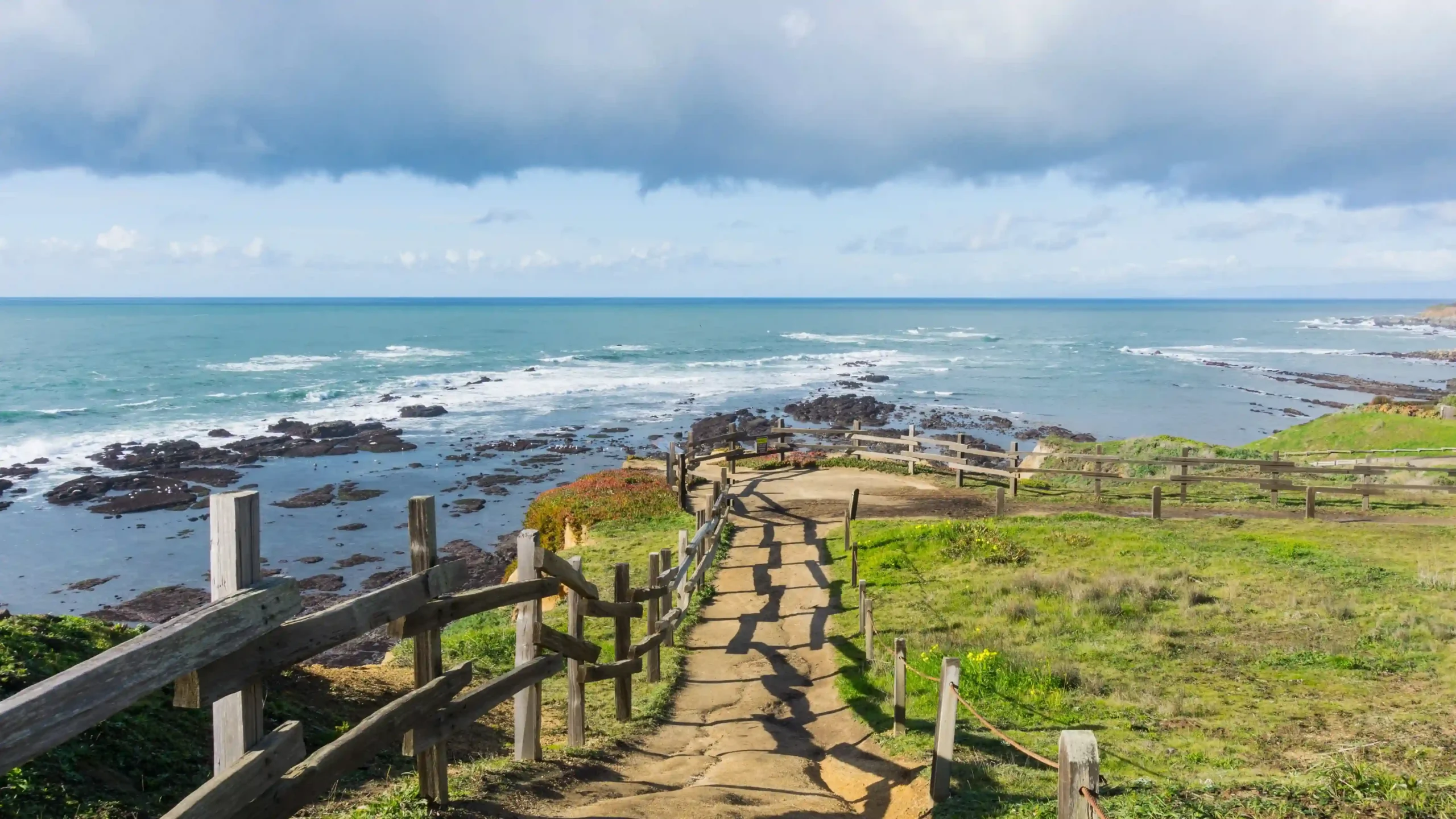 View of a rustic, fenced trail descending towards rocky ocean tide pools along a coastal bluff.