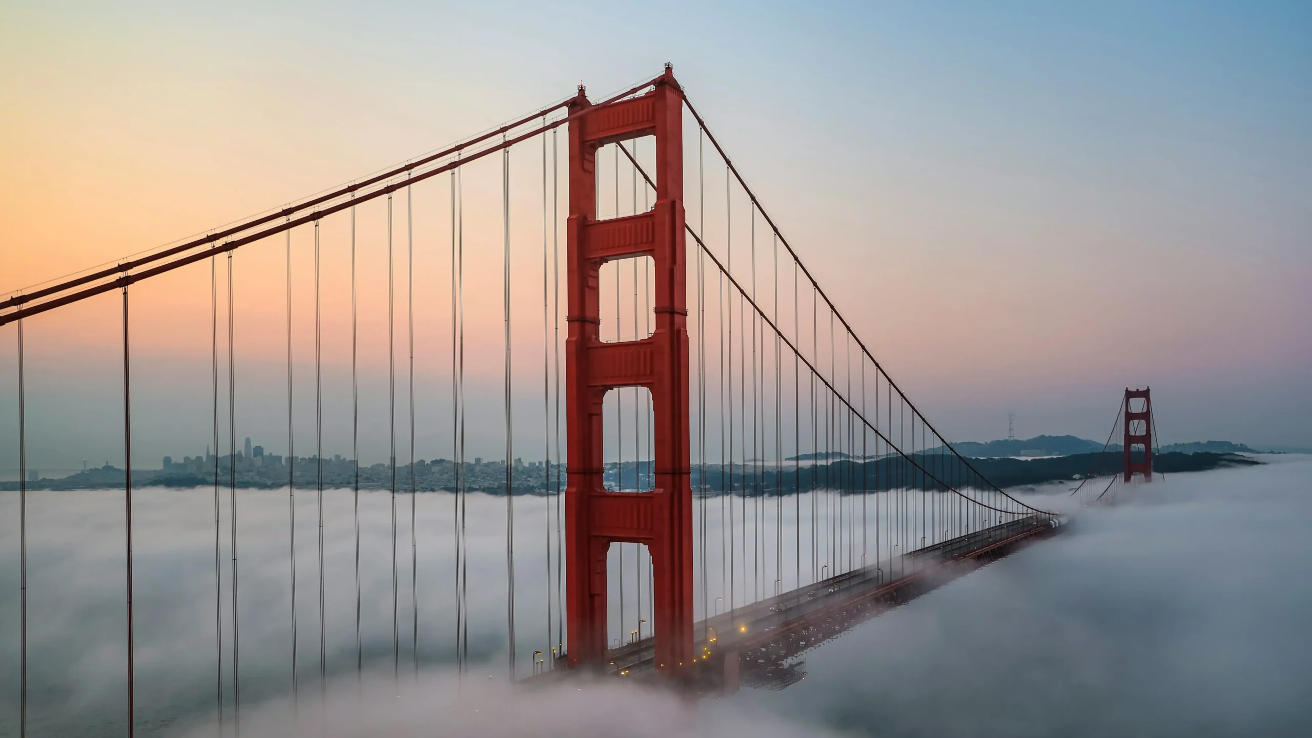 An aerial view of the iconic red towers of the Golden Gate Bridge standing high above a thick layer of white fog blanketing the bay.
