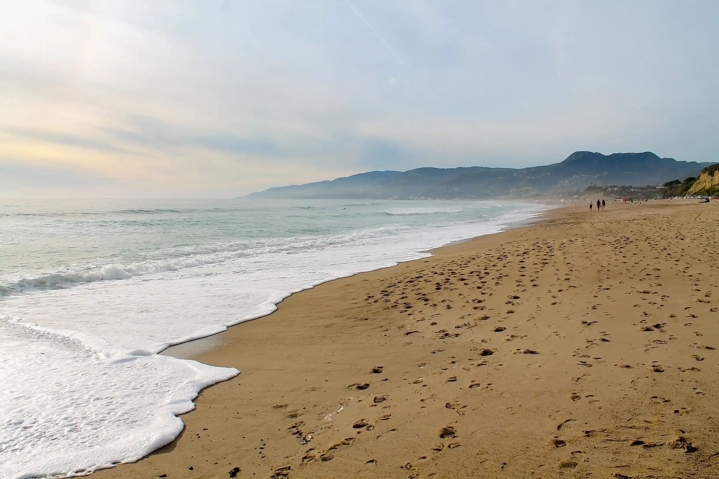 A wide, sandy beach stretching along the Pacific coastline with gentle white foam from waves washing ashore.