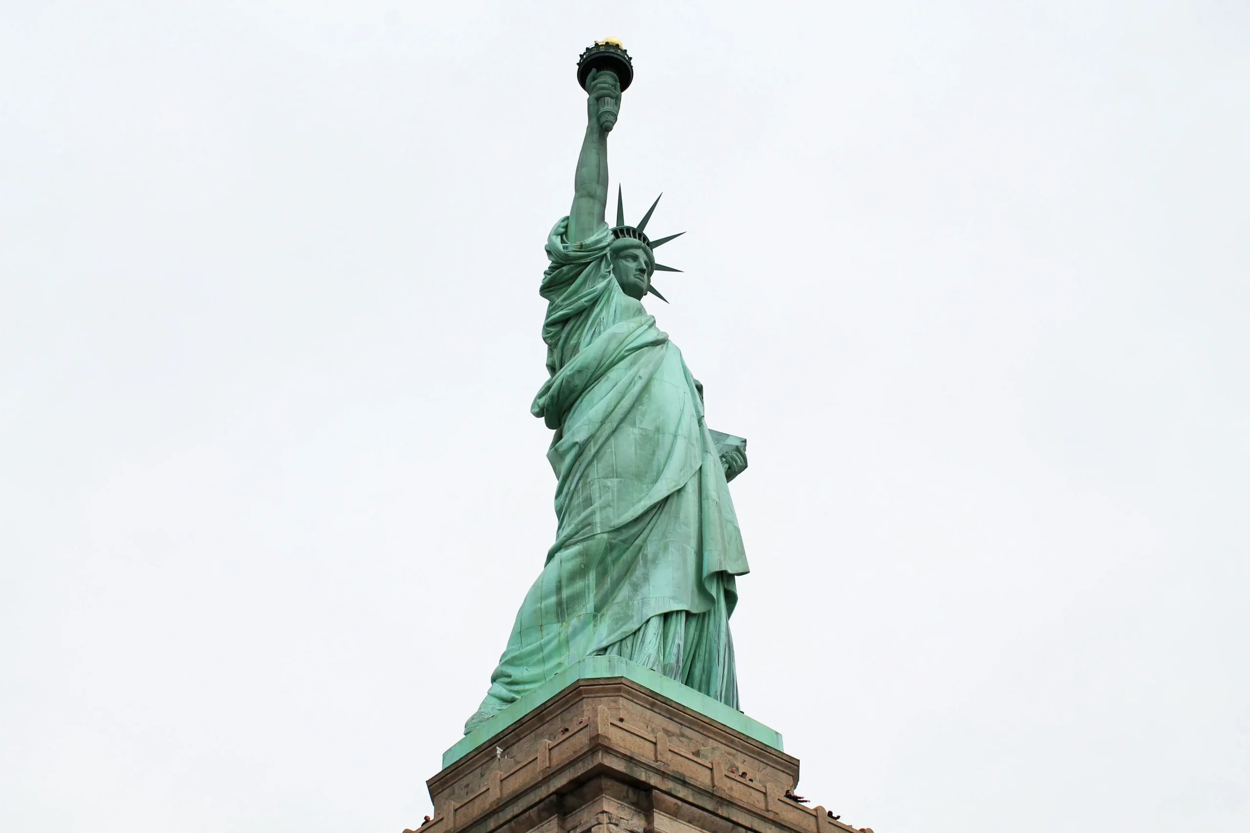 Shot of the Statue of Liberty looking up from its stone pedestal against a pale sky.