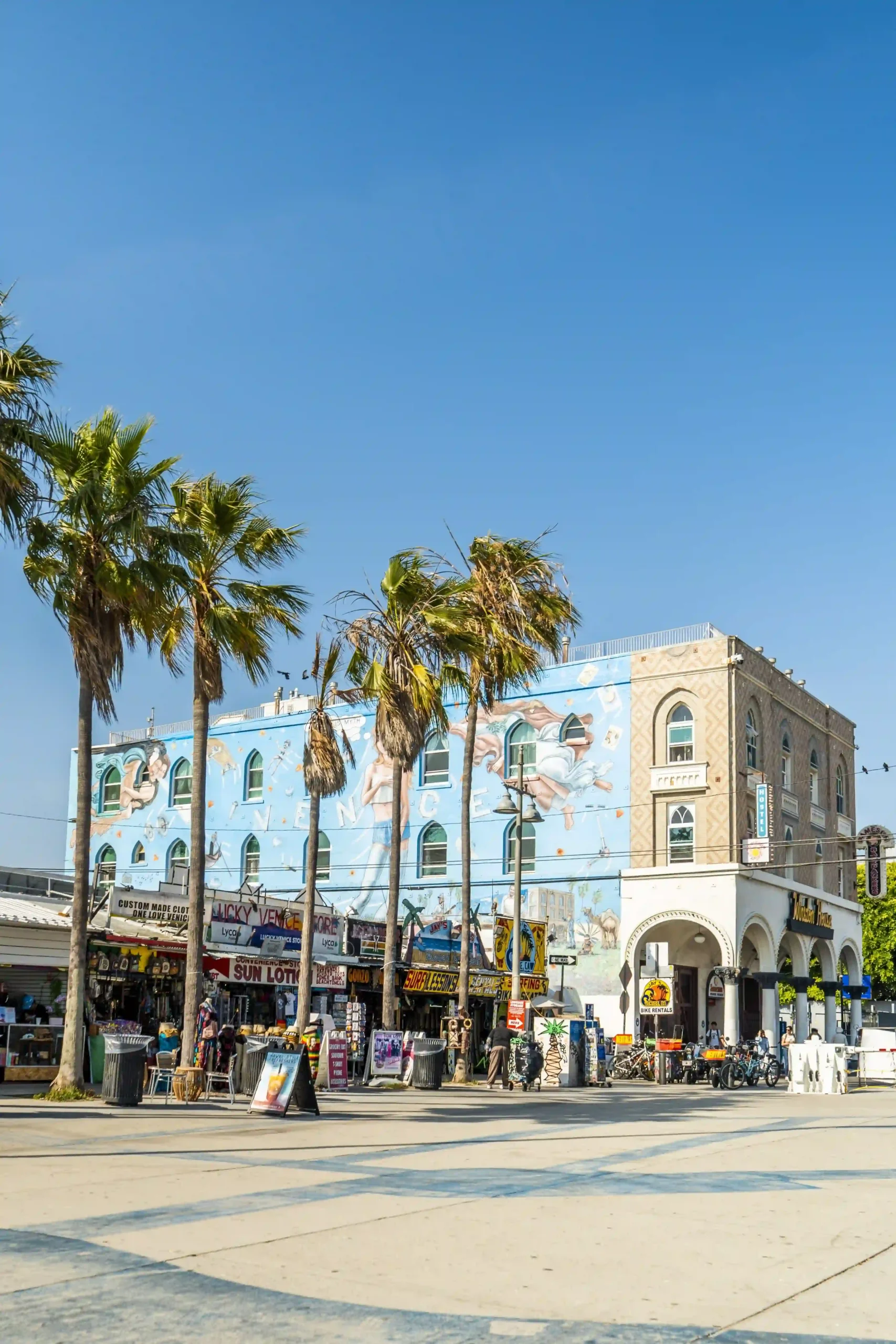 View of Venice Beach Boardwalk looking towards storefronts and a large building with a blue sky-themed mural featuring the word \