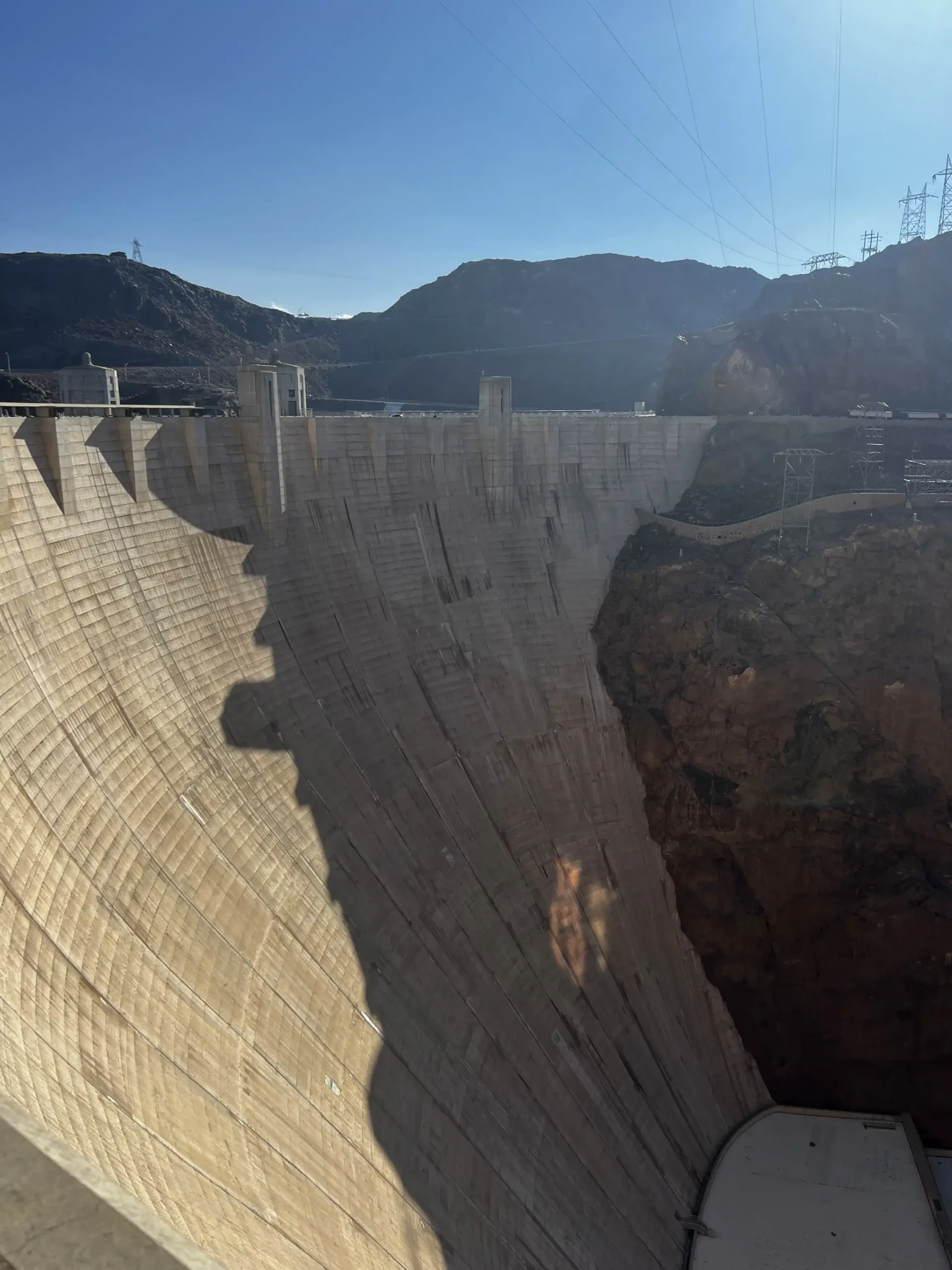 A wide-angle view looking down the massive concrete face of Hoover Dam, casting a large shadow over its curved structure, with the rocky cliffs of Black Canyon.