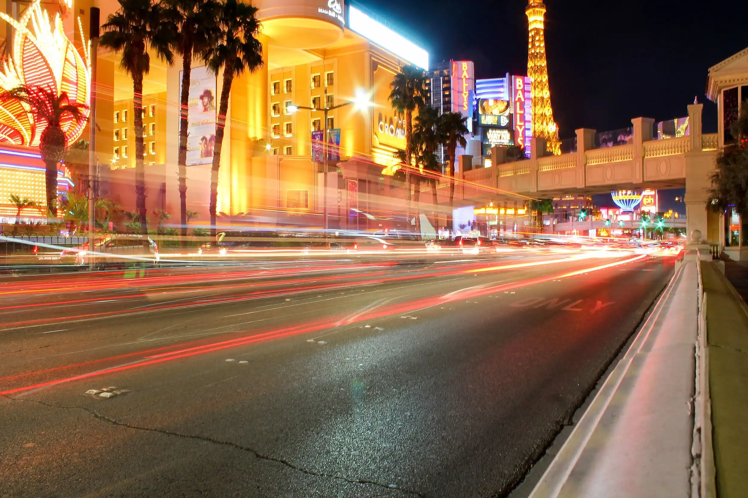 Las Vegas Strip at night, featuring motion-blurred streaks of red and white light from passing traffic against a backdrop of glowing hotels, palm trees, and the Eiffel Tower replica.