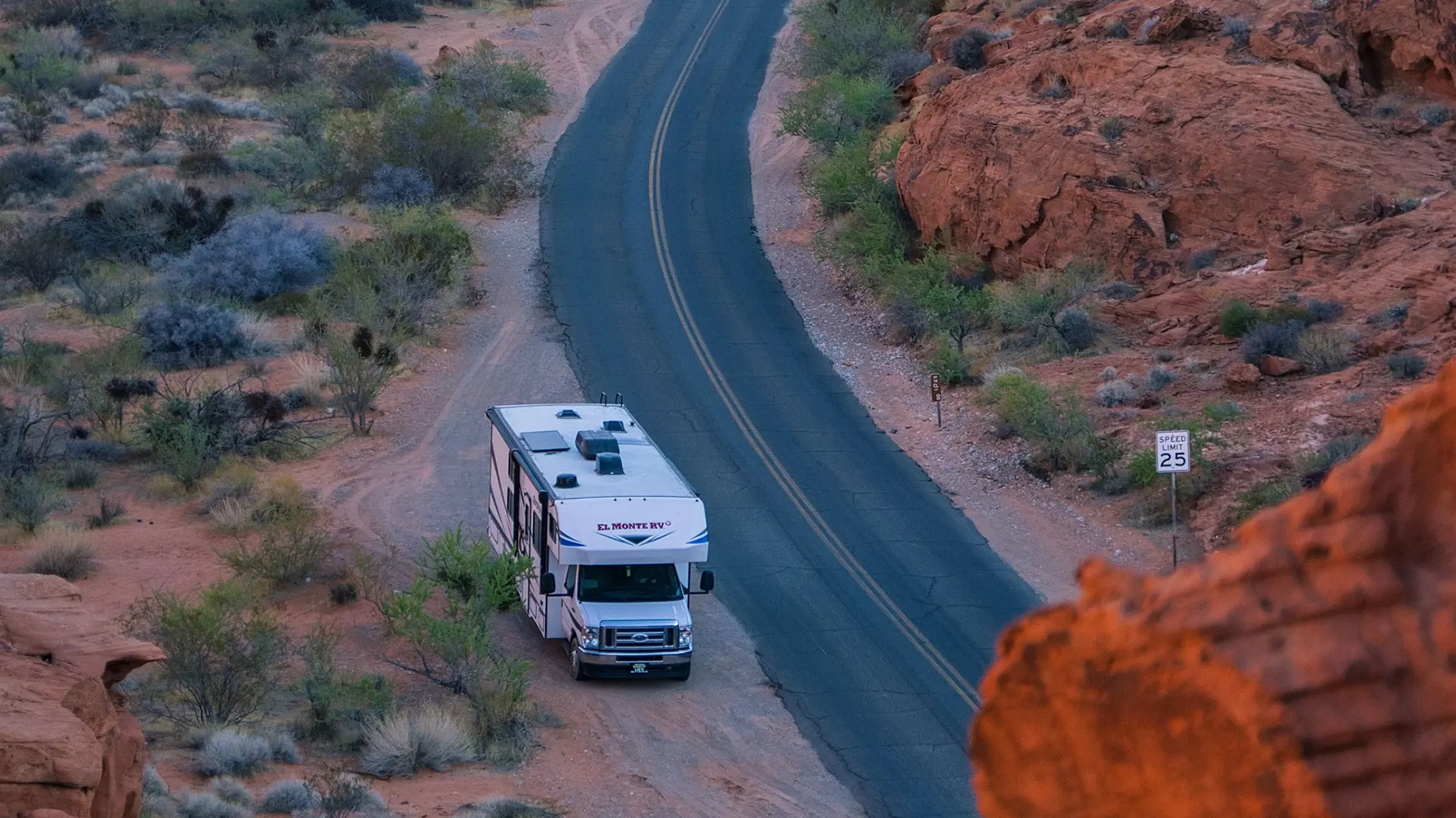 An overhead view of a white El Monte RV parked on the shoulder of a winding asphalt road cutting through a rugged desert canyon filled with red rock formations.