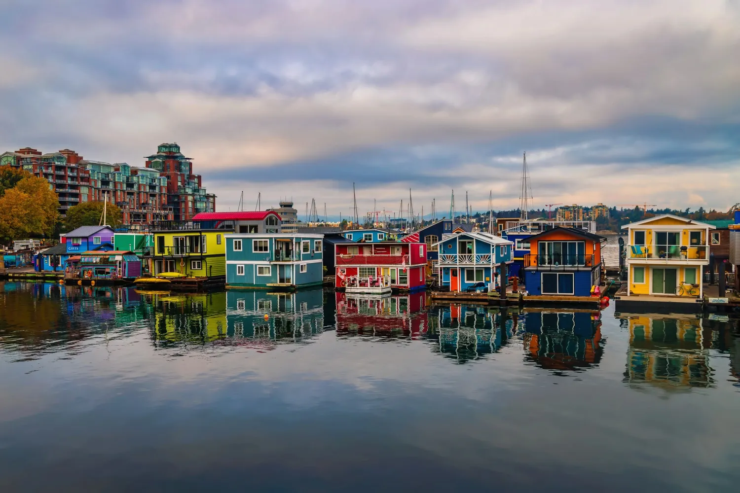Colorful houseboats at Fisherman’s Wharf reflected in the calm water under a cloudy sky.