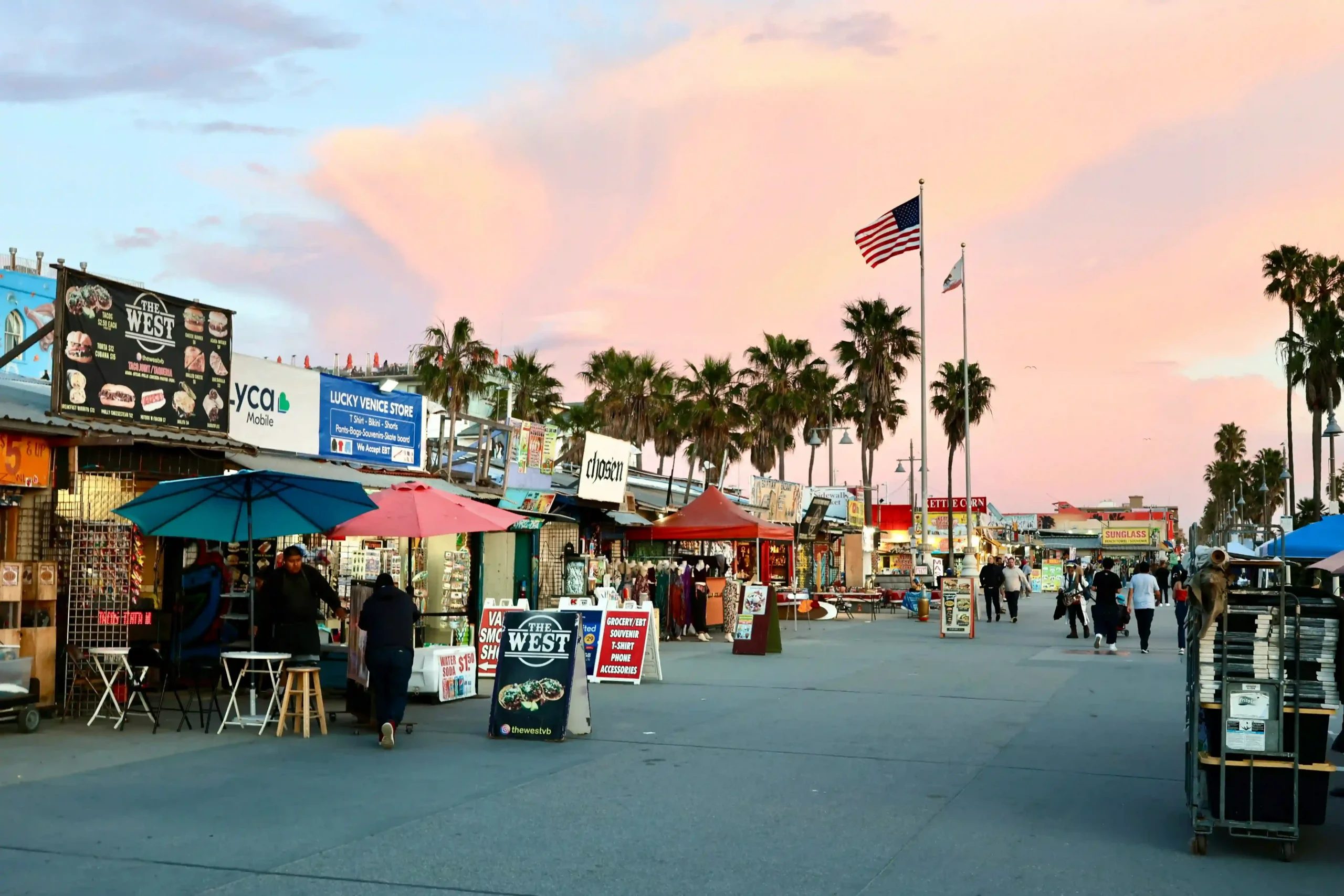 The Venice Beach Boardwalk at sunset, featuring colorful market stalls, palm trees, the American flag, and people walking along the oceanfront under a pink and orange sky.
