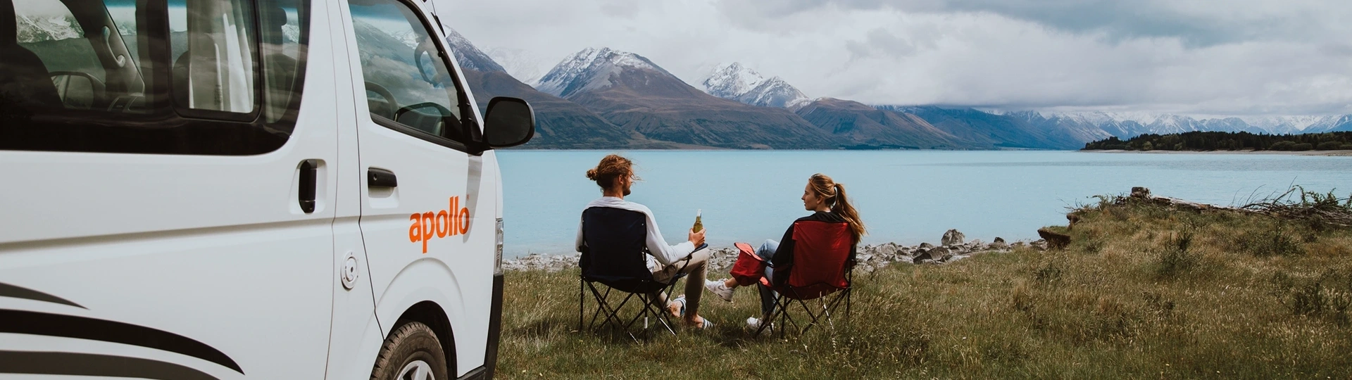 Un couple est assis au bord d\'un lac dans un camping en Nouvelle-Zélande à côté de leur véhicule Apollo.