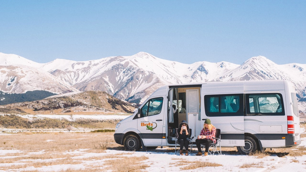 Un van est garé au bord de montagnes enneigées et deux personnes sont assises à côté du van.