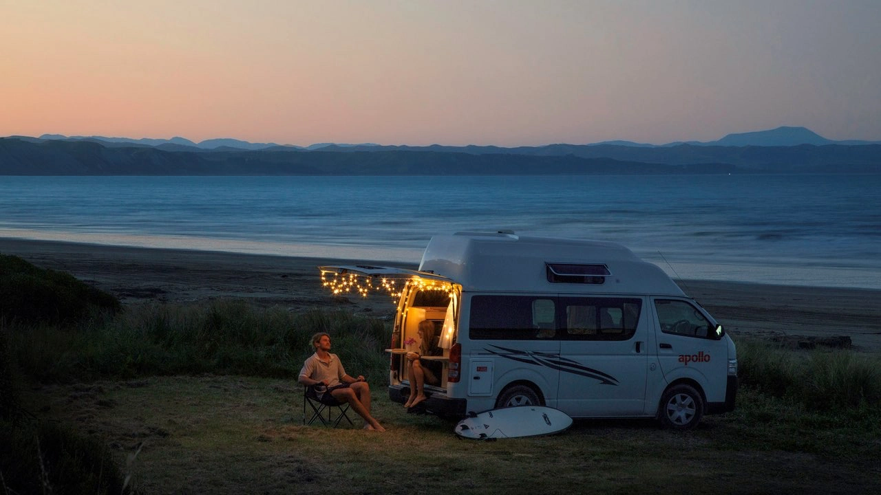 Un van est garé au bord d\'une plage sur la côte en Nouvelle-Zélande et deux personnes sont assises la nuit avec une guirlande lumineuse accrochée au van.